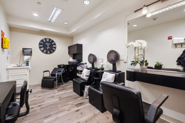 A hair salon with two dryer chairs, a styling chair, a large mirror, and a desk with a chair.