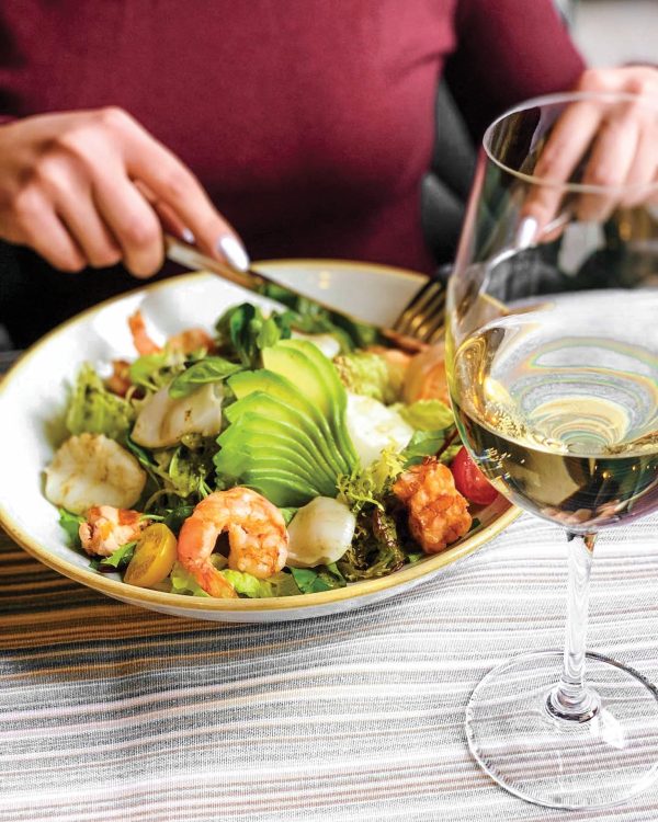 A closeup of a person eating a salad with avocado and shrimp, with a glass of wine to the side