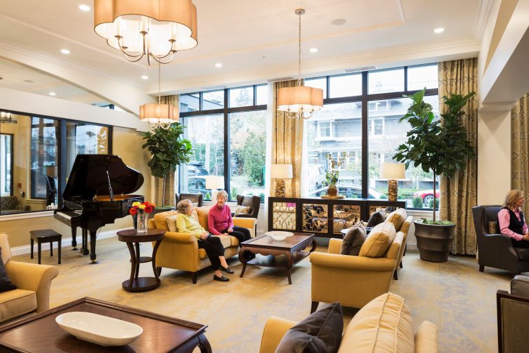 The lobby, with residents sitting and chatting in the middle, a piano to the left, and large windows at the back
