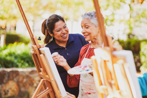 A smiling employee assists an elderly resident with her painting at an outdoor easel in a garden setting